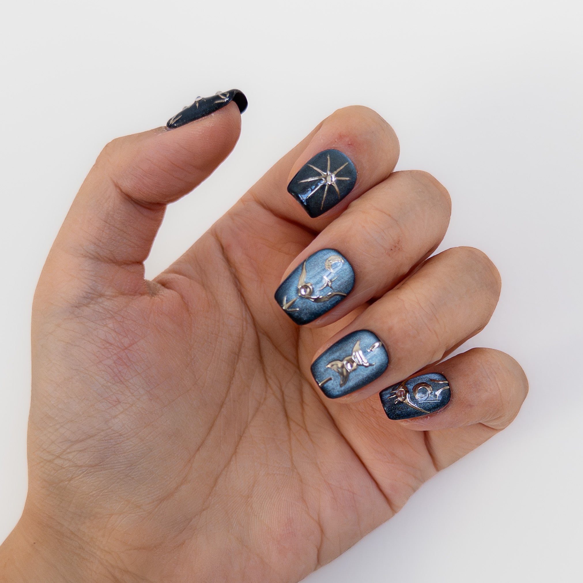 Close up of Astral Vault press on nails on a hand. Deep teal blue cat eye gel with raised silver chrome abstract stars and moons. Handcrafted short square manicure.