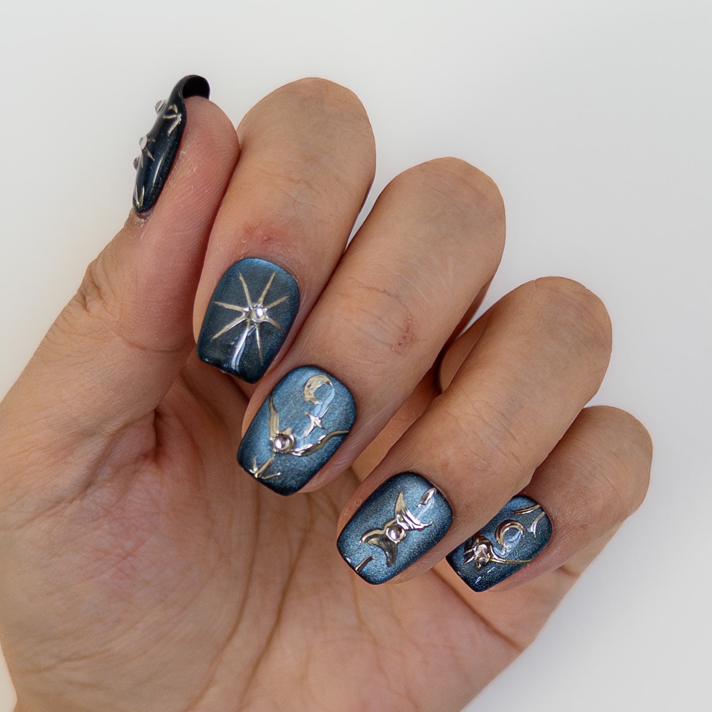 Angled view of Astral Vault handmade nails highlighting texture. Shimmering blue cat eye polish with metallic silver symbols and crystal accents. Reusable gel press ons.