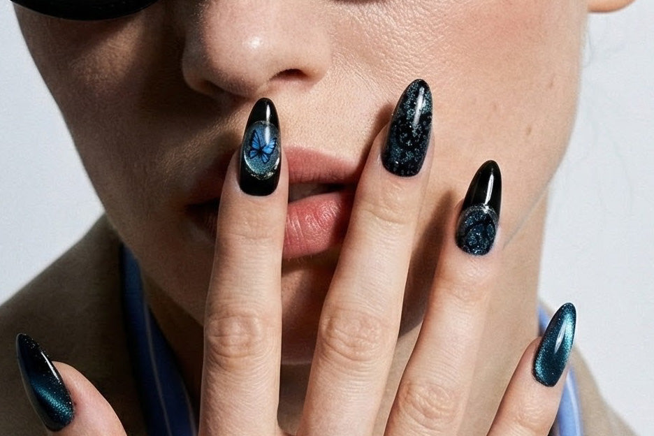 Close-up of a model's hand resting near the lips, displaying almond-shaped nails with a dark teal magnetic cat-eye finish and a central blue butterfly motif encapsulated on the accent nail.