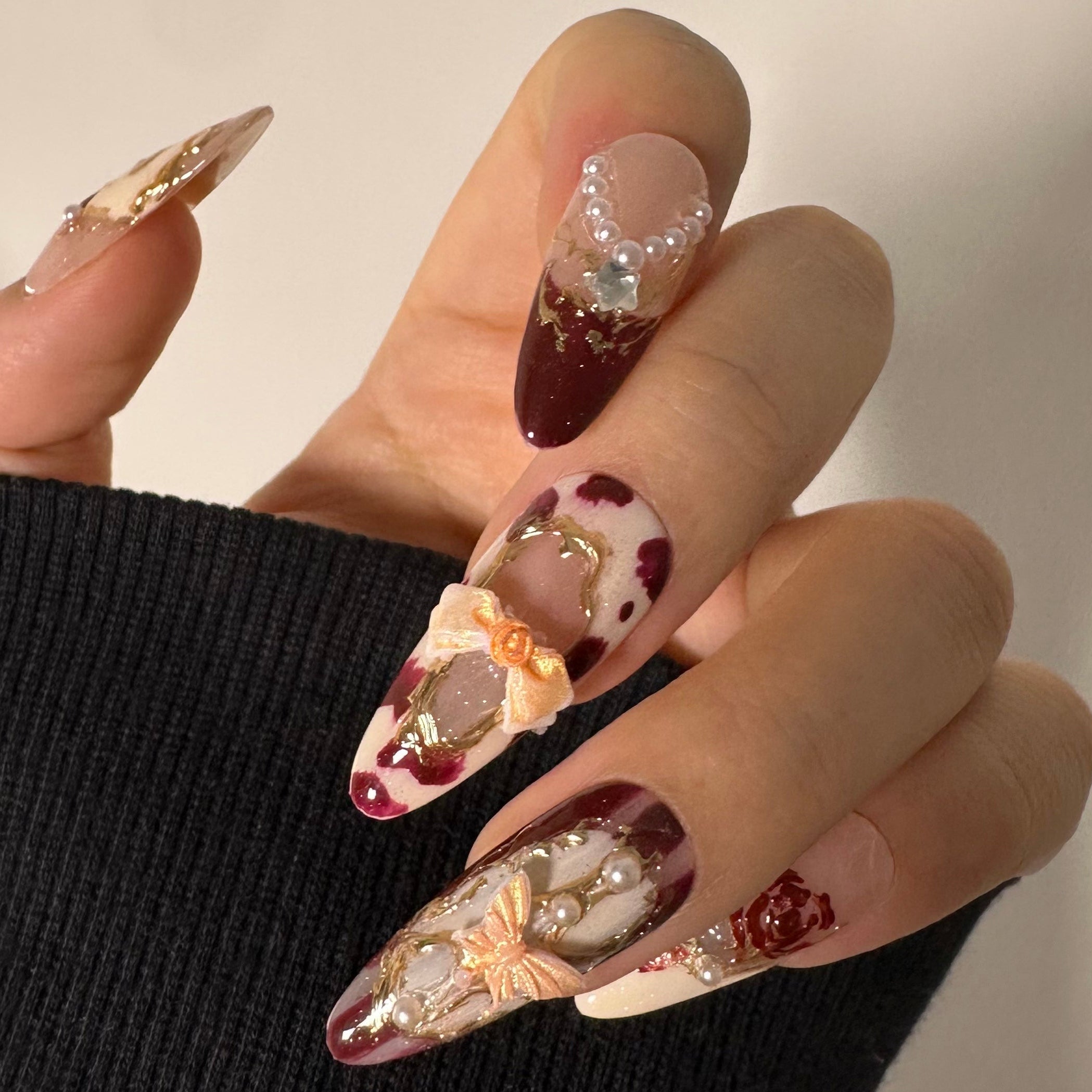 Close-up of 3D pearl chains and gold bow charms on wine red almond nails, featuring hand-painted rose art on salon-grade structured gel.