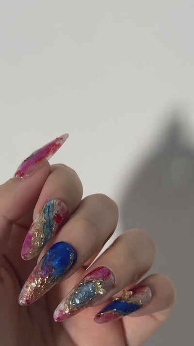 Close-up video of long stiletto press on nails featuring a dramatic abstract marble design. The nail art blends deep royal blue and magenta swirls on a translucent base, accentuated with luxury gold foil flakes for a textured, artistic finish.
