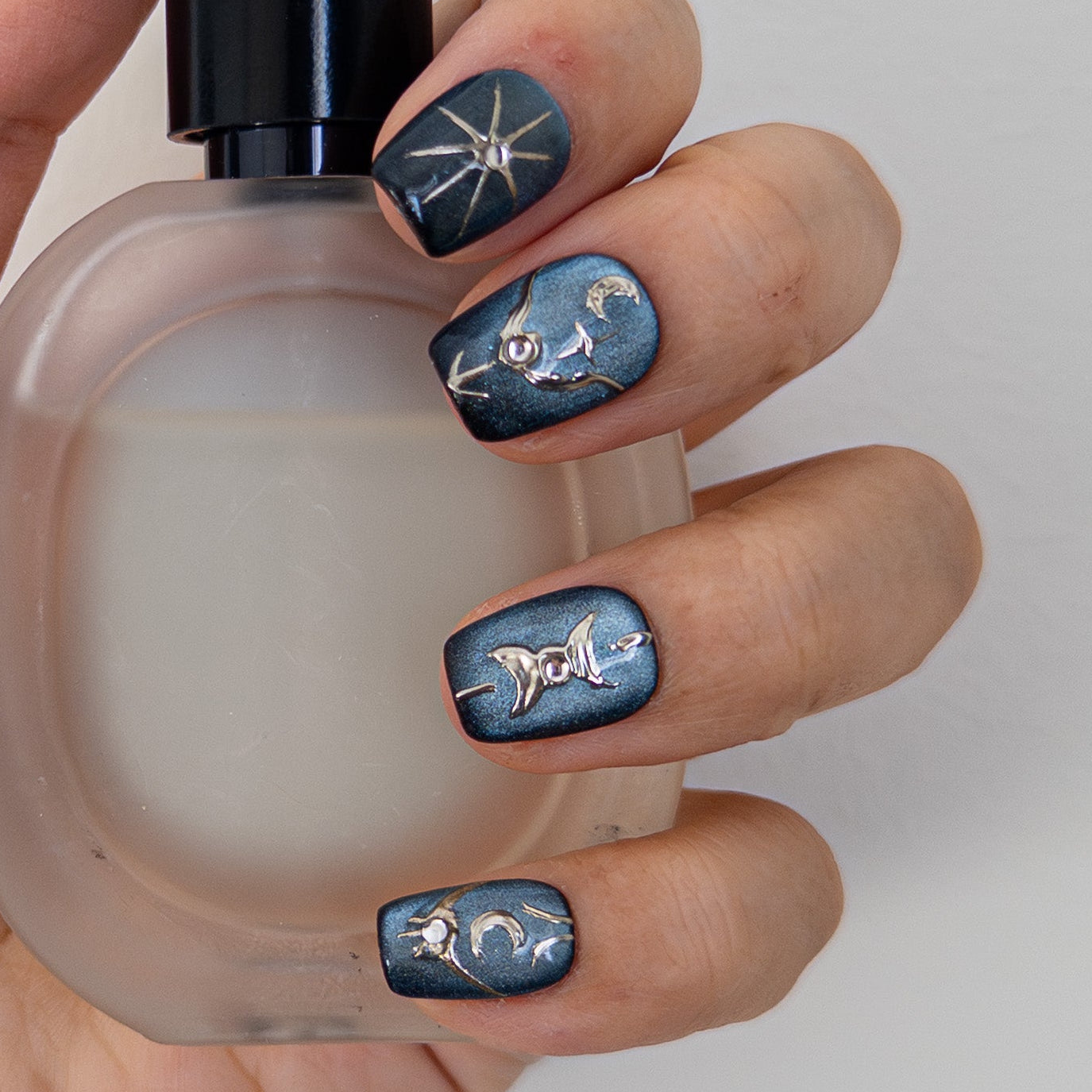 Astral Vault luxury press on nails holding a perfume bottle. Dark blue magnetic cat eye finish with 3D silver chrome celestial designs and rhinestones. Short square shape.