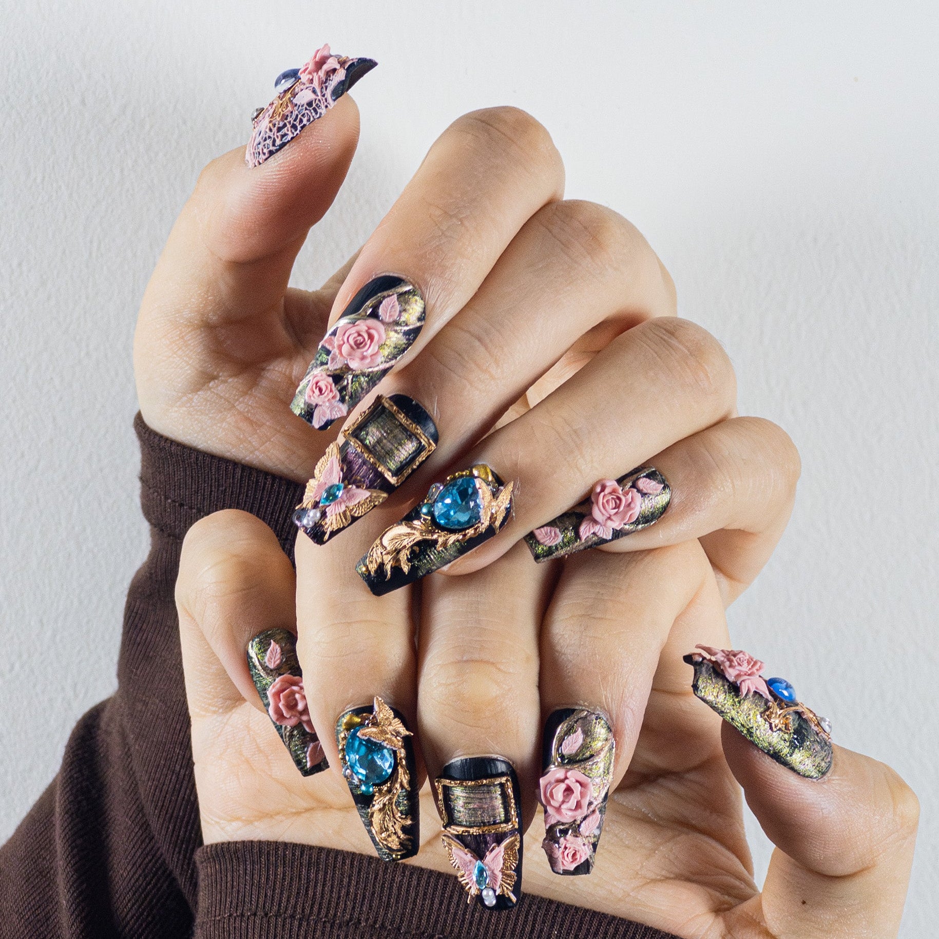 Hands wearing elaborate, long coffin-shaped press-on nails. The design is a dark, gothic floral theme with a textured, dark background that has gold and purple highlights. There are 3D sculpted pink roses, green leaves, and various blue and teal gems, along with gold rectangular frames on some nails.