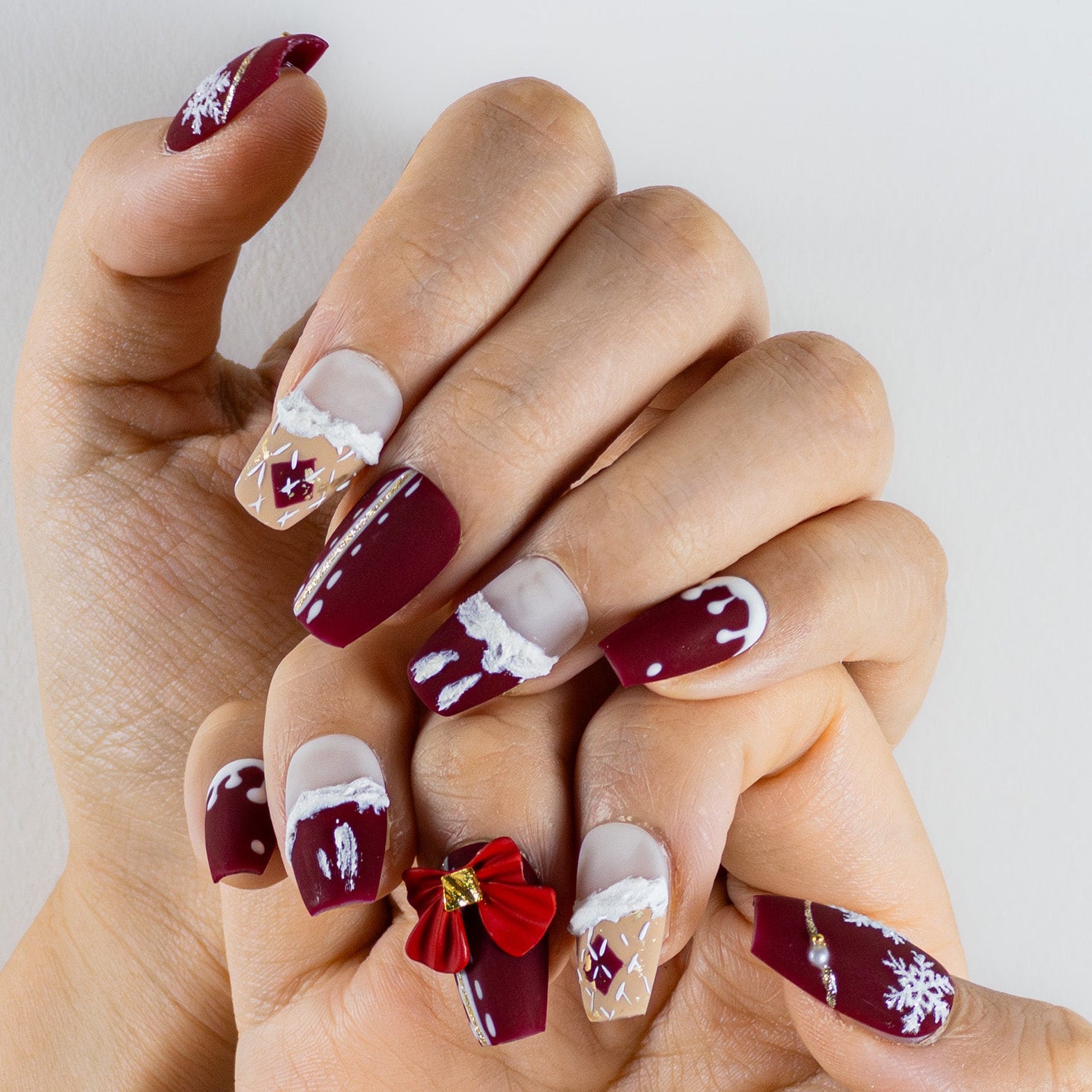 A close-up of hands wearing short, square-shaped press-on nails with a matte finish. The primary colors are deep red and beige. The design includes hand-painted white snowflake patterns, white textured 'snow' drips on the tips, and a large, red 3D bow charm on one ring finger.