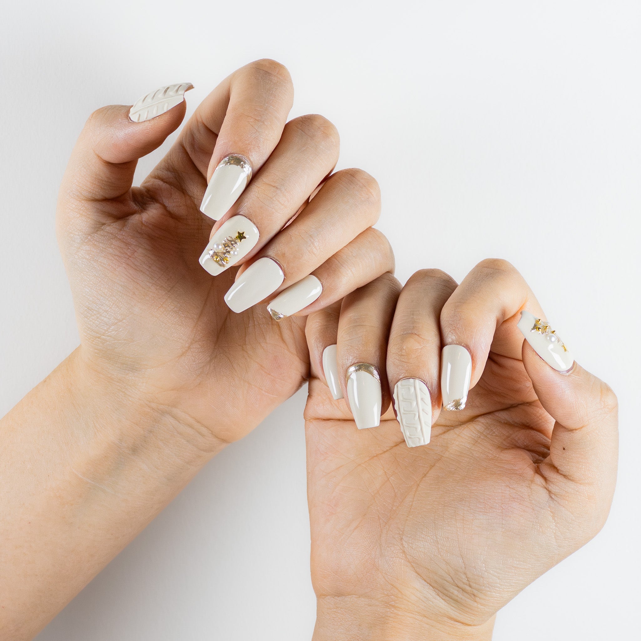 Two hands with long, coffin-shaped press-on nails are shown. The nails are a glossy white color. One thumbnail has a textured, cable-knit sweater pattern. The ring fingers feature Christmas tree designs made with small gold studs, pearls, and a star charm at the top. Other nails have french tip outlines in gold glitter.