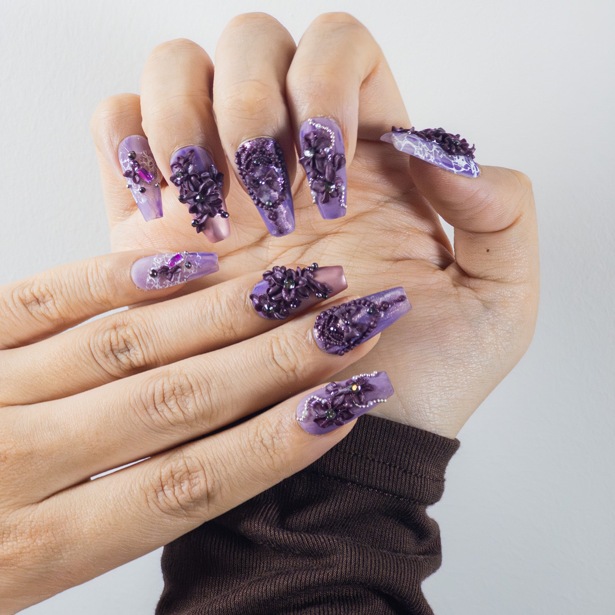 Hands display long, coffin-shaped press-on nails in a purple monochromatic theme. The nails have a matte finish with various textures. Features include 3D sculpted purple flowers and leaves, clusters of purple and clear gems, small pearls, and some nails with a lace-like pattern.