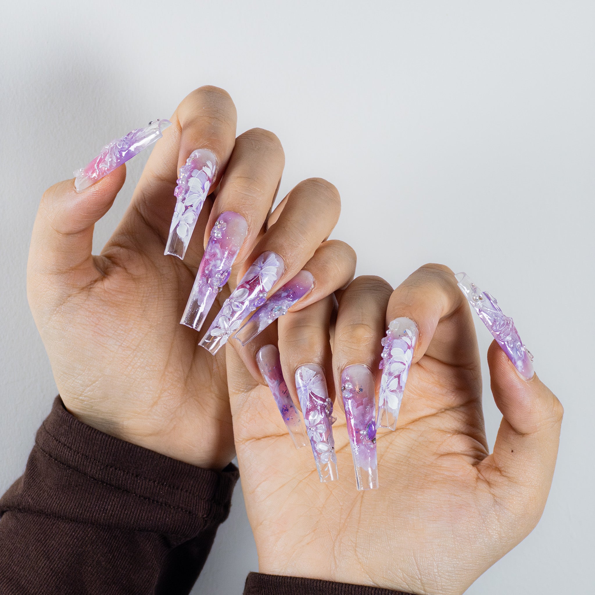Hands holding a pose to show off extra-long, clear stiletto press-on nails. The nails have a translucent, purple and white marble or floral design. They are highly decorated with white 3D flowers, small pearls, and scattered glitter, giving them an icy or crystalline appearance.